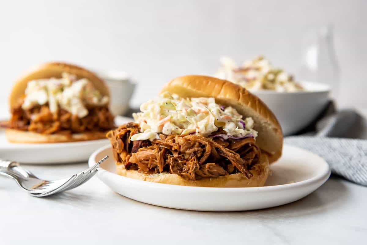 Two apricot pulled pork sandwiches on plates in front of a bowl of coleslaw.