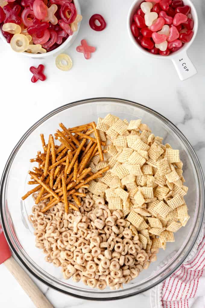 Adding pretzel sticks, Chex cereal, and cheerios to a bowl of melted white chocolate.