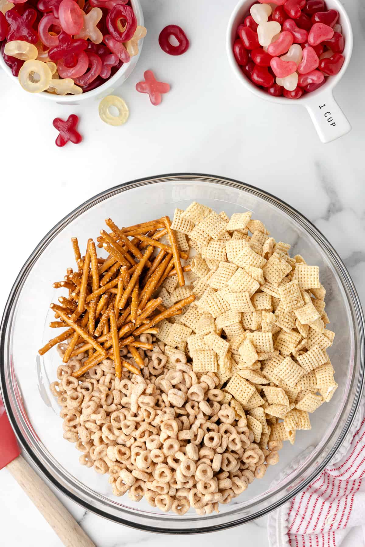 Adding pretzel sticks, Chex cereal, and cheerios to a bowl of melted white chocolate.