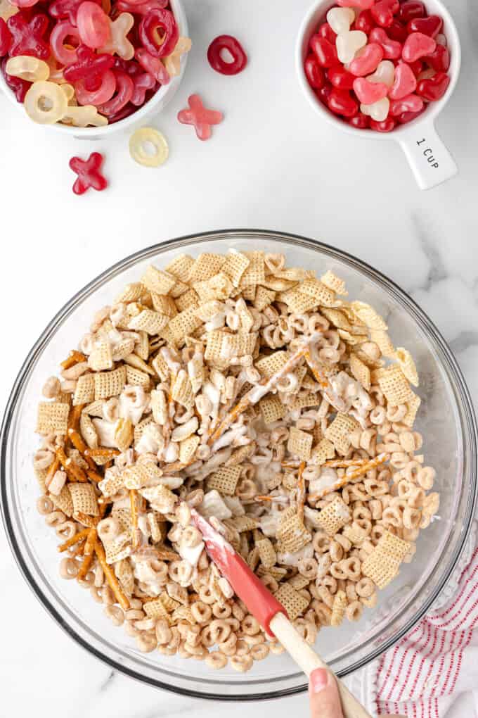 Mixing up cereal, pretzels, and melted white chocolate in a large glass bo with a spatula.