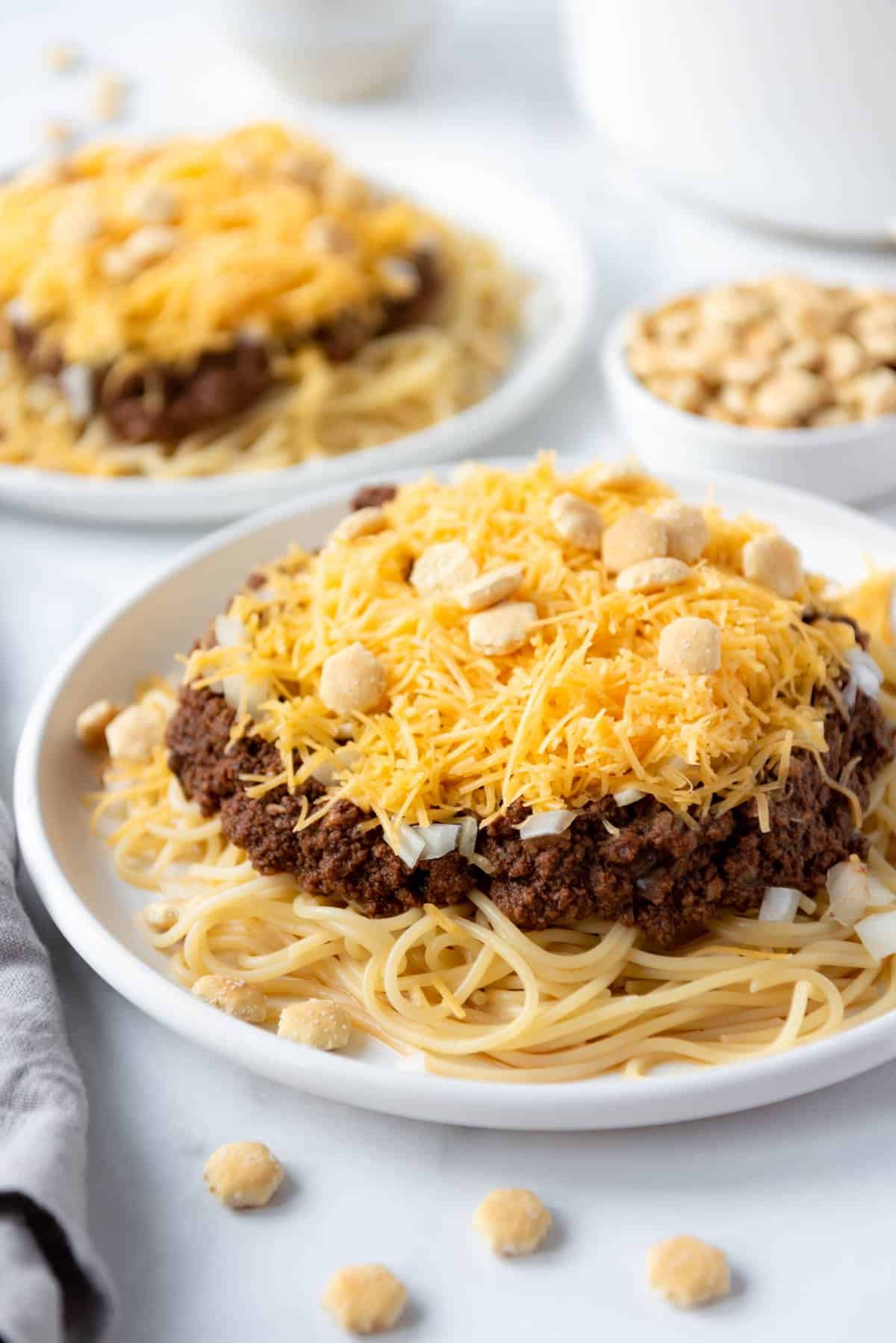 Two white plates with spaghetti and Cincinnati Chili topped with cheese and oyster crackers.