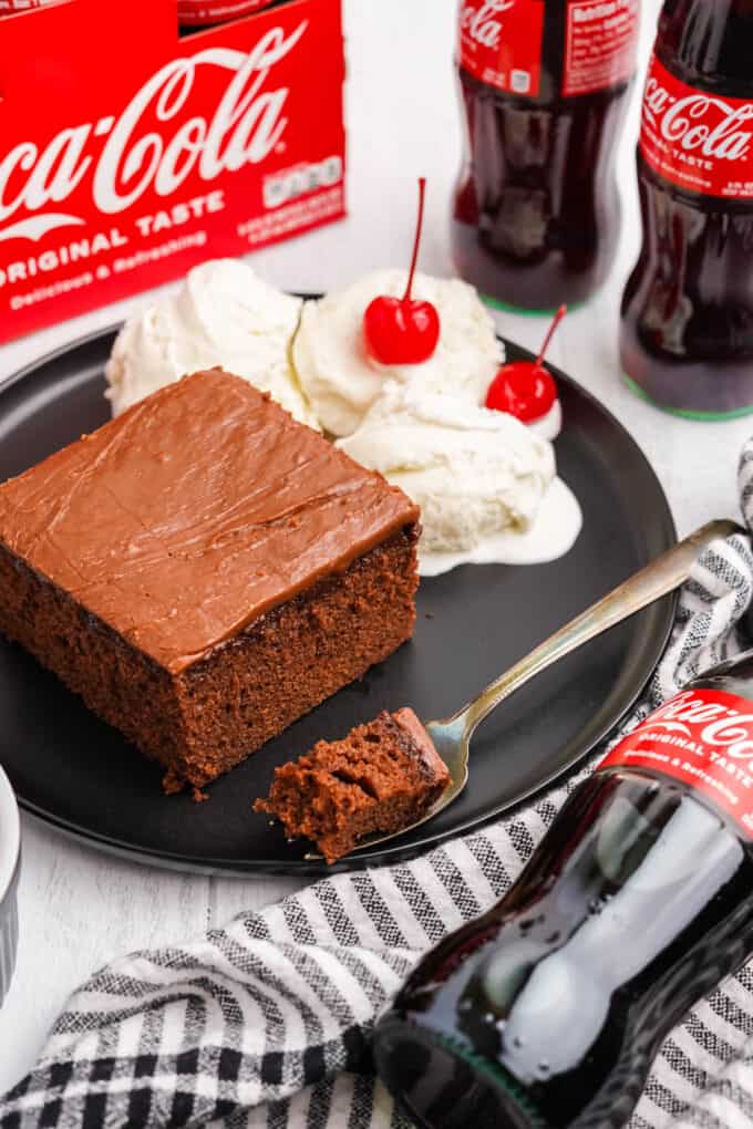 A piece of coca cola cake on a plate with ice cream and cherries next to bottles of coca cola soda.