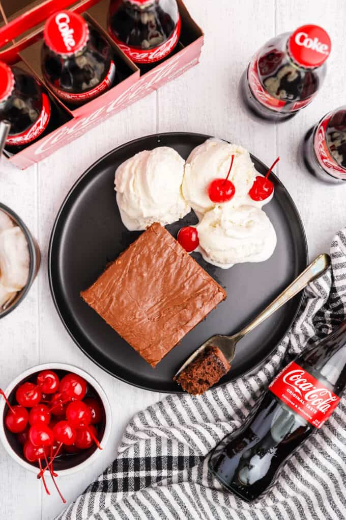 A black plate with a square of coca cola cake, ice cream, and maraschino cherries.