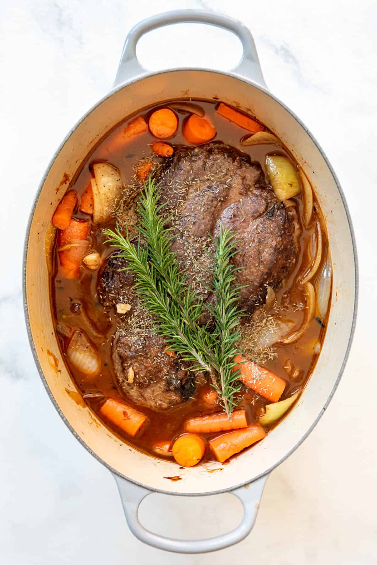 Adding rosemary sprigs and beef broth to a beef pot roast, carrots, and onions in a Dutch oven.
