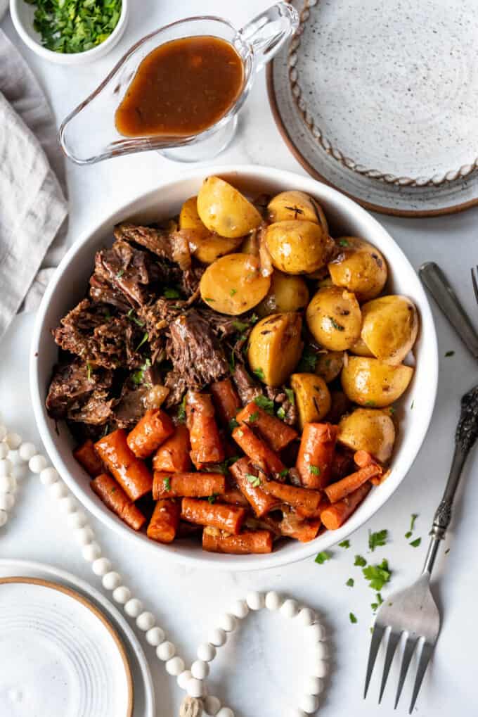 A large white serving bowl of pot roast, potatoes, and carrots next to plates and gravy in a glass dish.