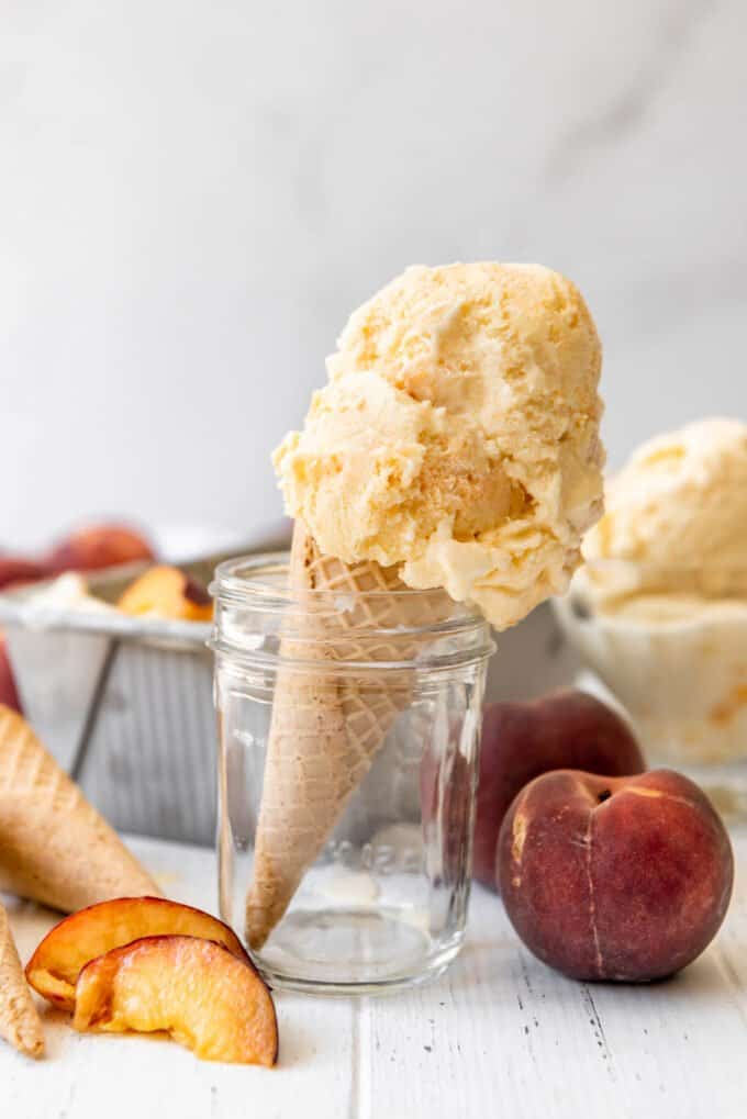 An sugar cone with a scoop of peach ice cream standing in a glass jar next to sliced fresh peaches.