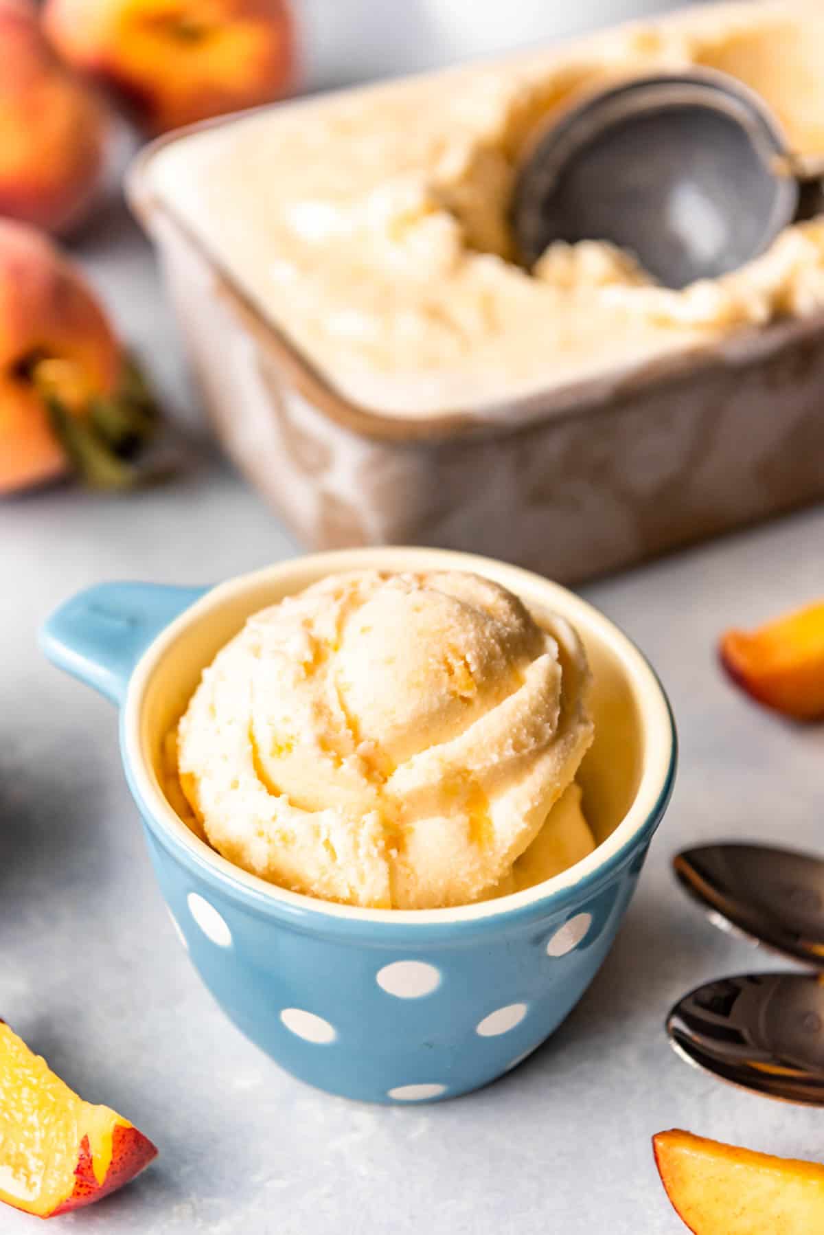 Fresh peach ice cream in a loaf pan with a scooper and a scoop in a cup with fresh peaches around it.