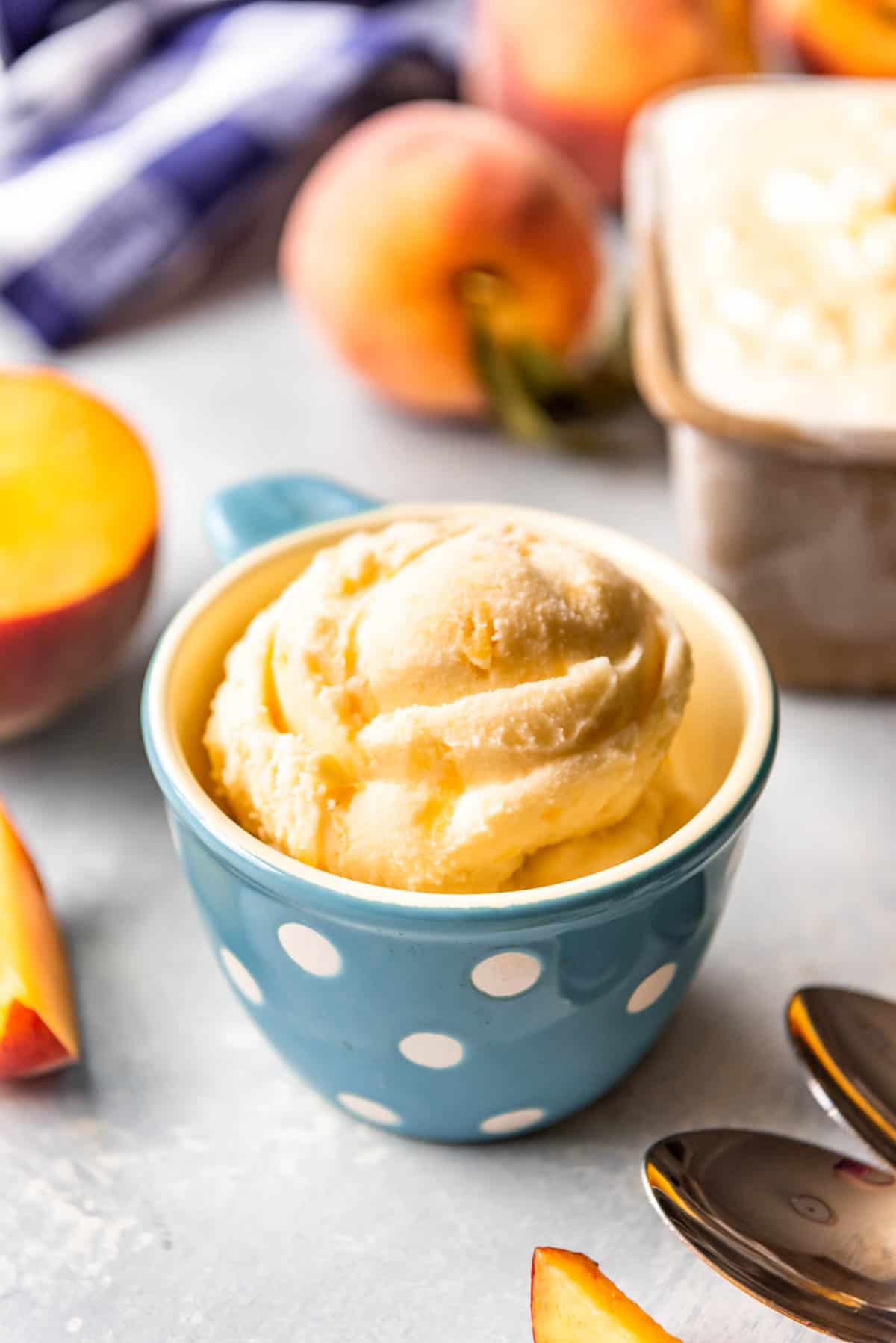 A cup with a scoop of peach ice cream inside and fresh peaches behind it.