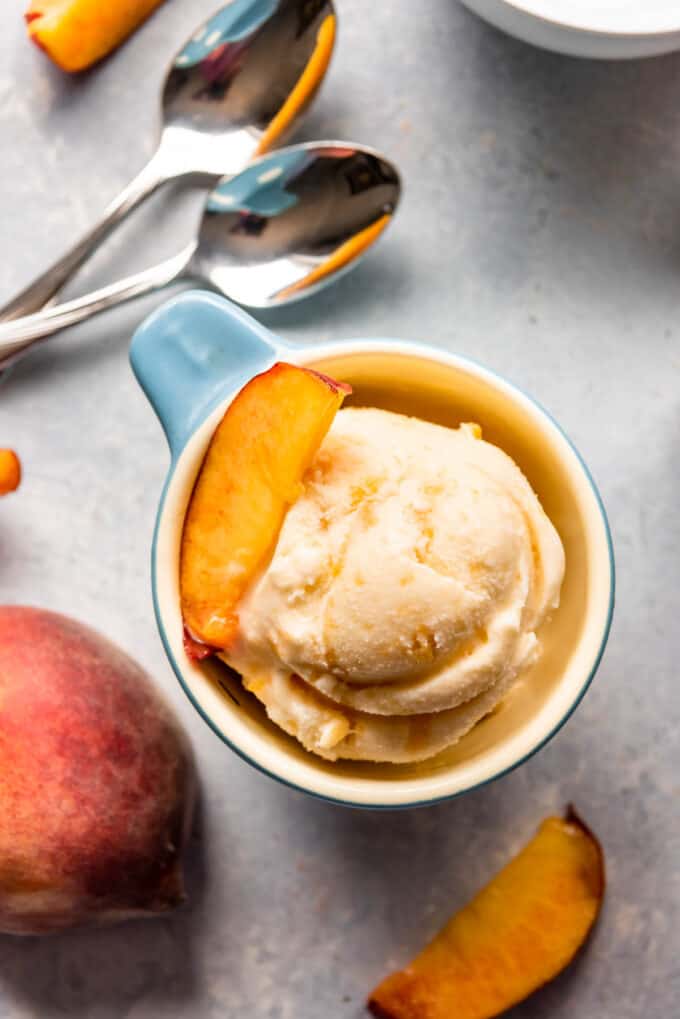A scoop of peach ice cream in a small bowl next to spoons with a slice of peach on top.