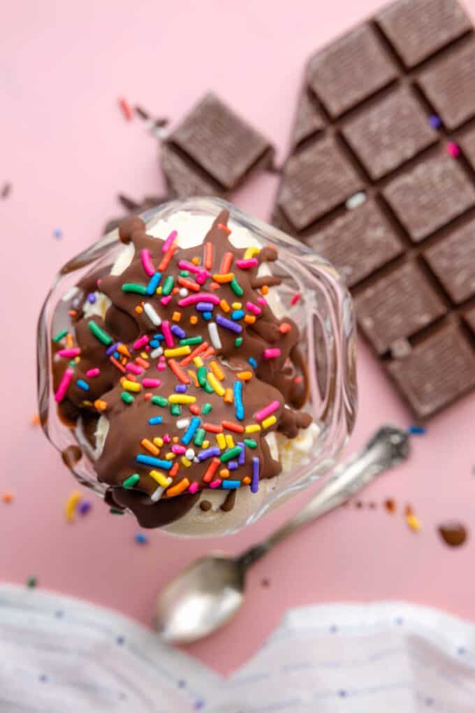 Hardened chocolate magic shell topping with sprinkles over scoops of vanilla ice cream in a bowl next to chocolate bars.