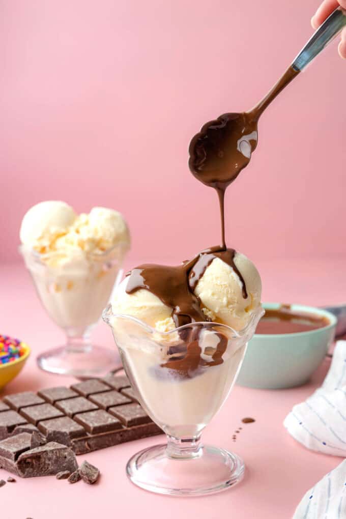 A spoon drizzling homemade chocolate magic shell onto scoops of vanilla ice cream in glass cups.