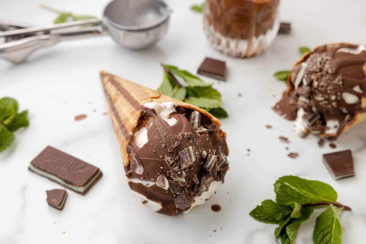 An ice cream cone with mint chocolate magic shell topping next to fresh mint leaves.