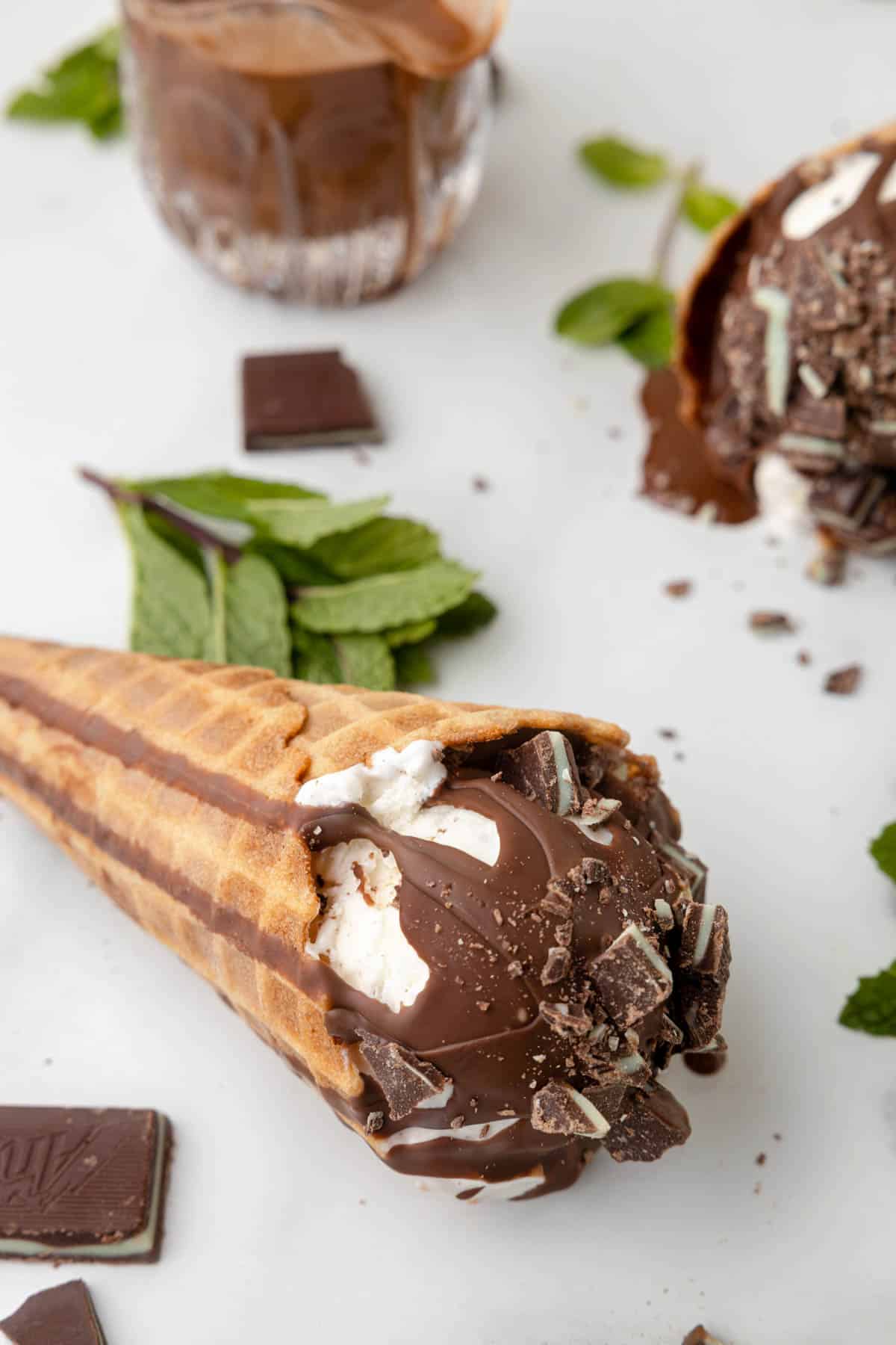 An ice cream cone with mint chocolate magic shell topping next to fresh mint leaves.