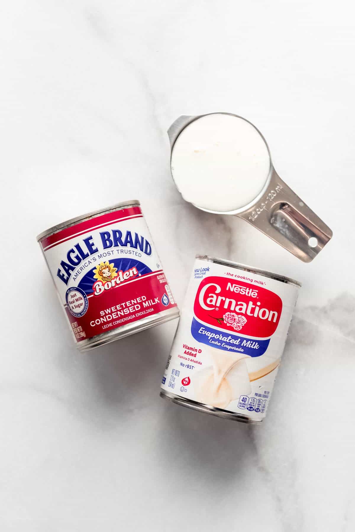 Cans of evaporated milk, sweetened condensed milk, and whole milk in a measuring cup.