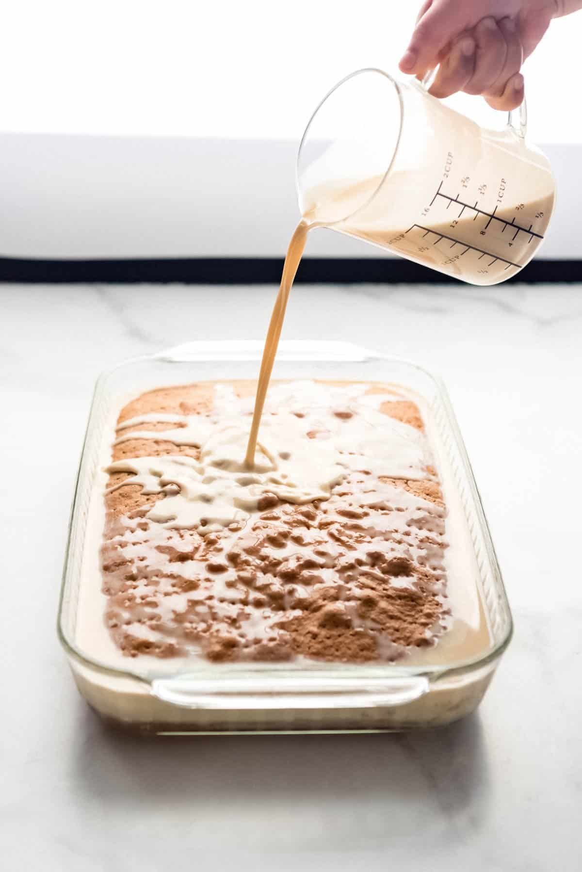 A hand holding a glass pitcher of sweet milk being poured over a cake.