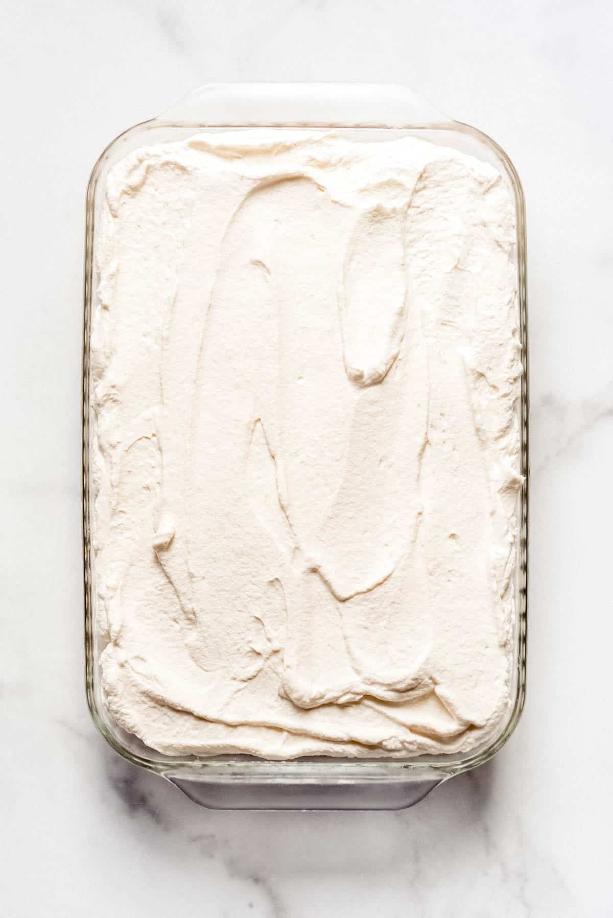 Homemade whipped cream spread over tres leches cake in a glass rectangular baking dish.