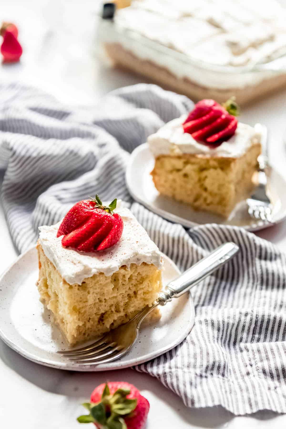 Two slices of tres leches cake on serving plates with forks and topped with fanned strawberries.