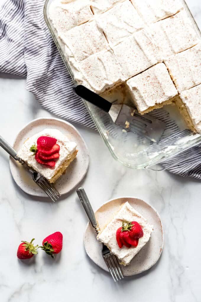 An overhead image of sliced tres leches cake on white plates with strawberries and in a baking dish sliced and ready to serve.