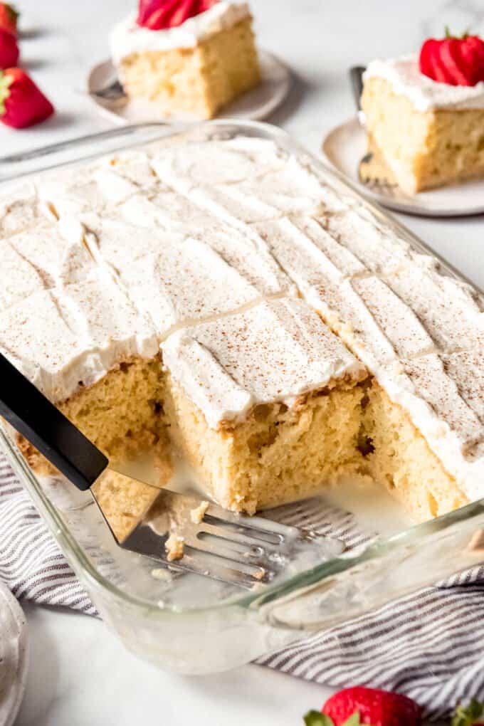 A baking dish with sliced and frosted tres leches cake with some slices removed.