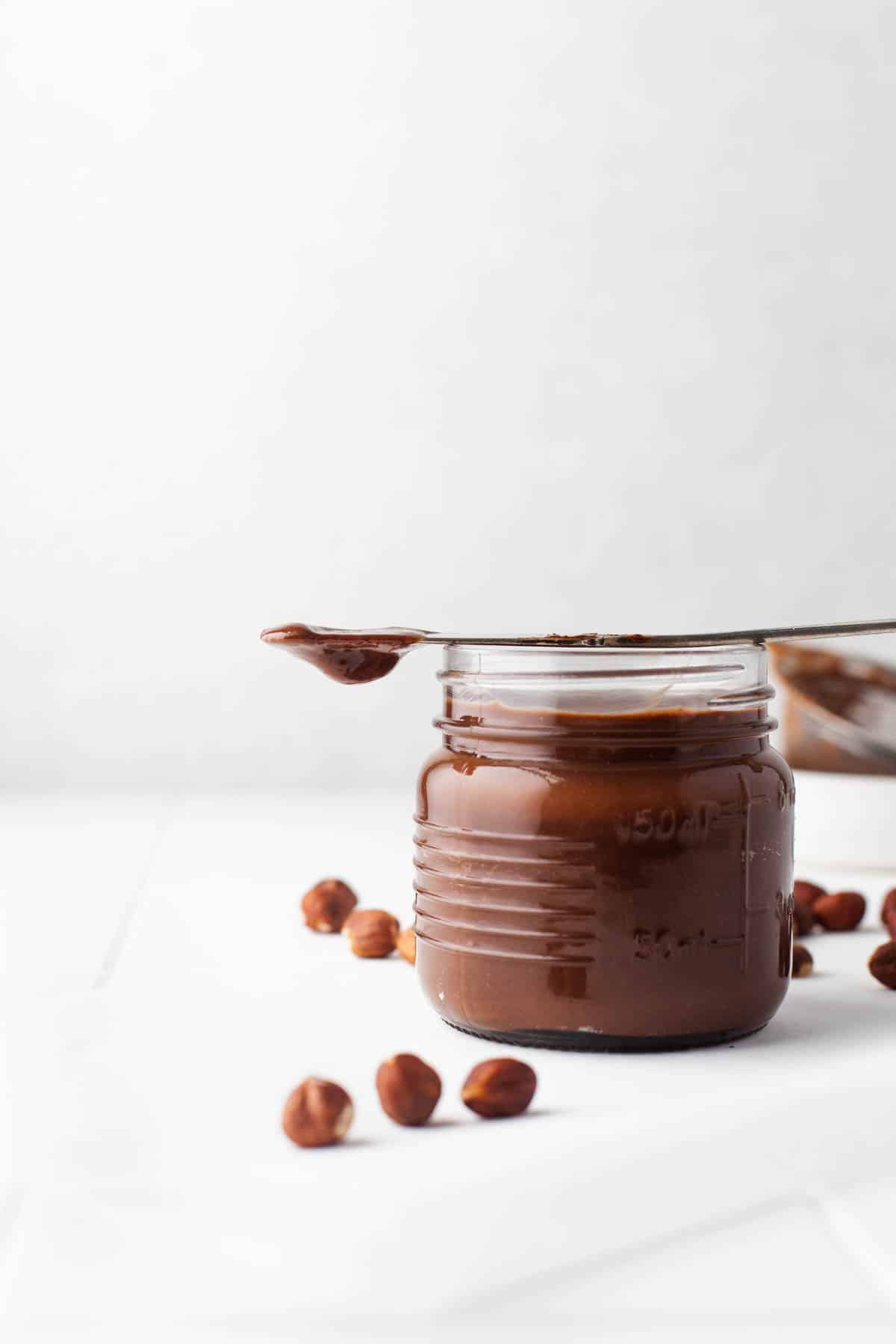 A knife dripping homemade Nutella set on top of a jar of the creamy spread.