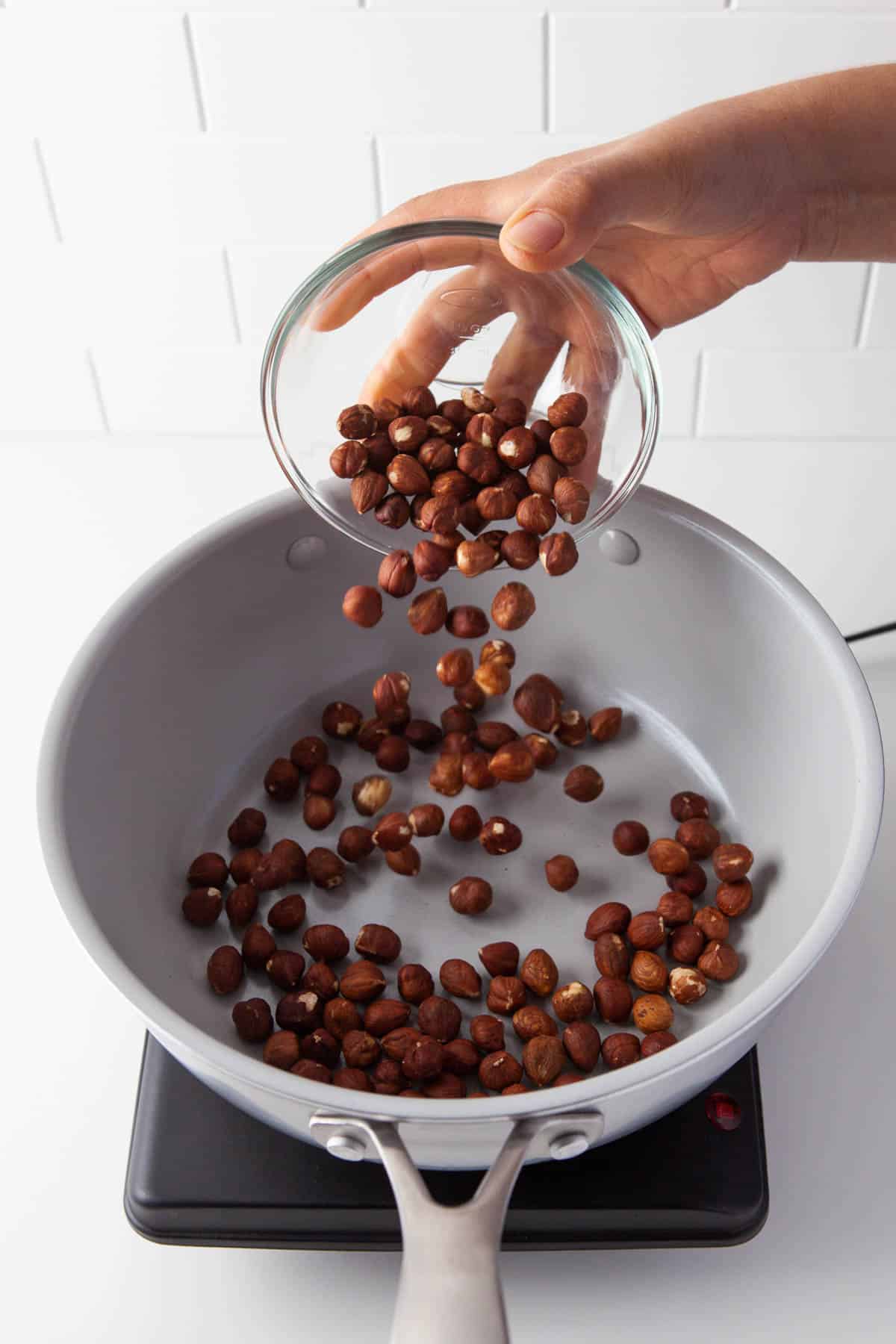 A hand pouring hazelnuts into a skillet from a glass bowl.