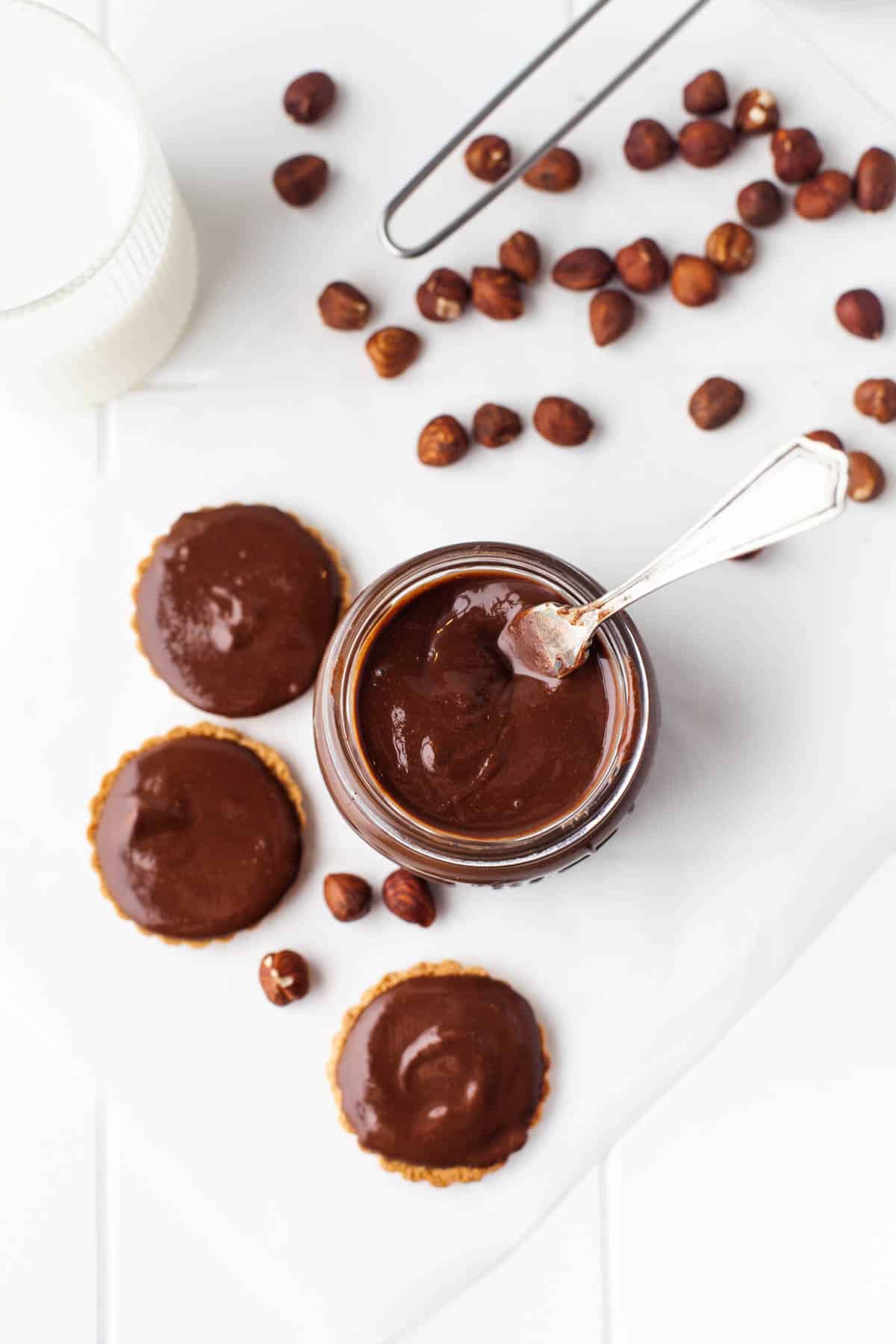 A glossy, smooth homemade chocolate hazelnut spread in a jar and on cookies next to hazelnuts.