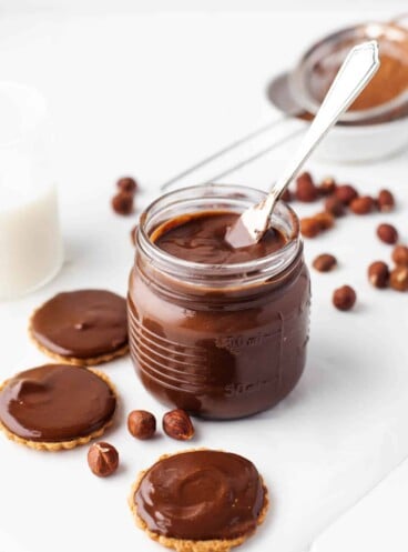 Homemade Nutella in a glass jar with cookies and hazelnuts surrounding it.