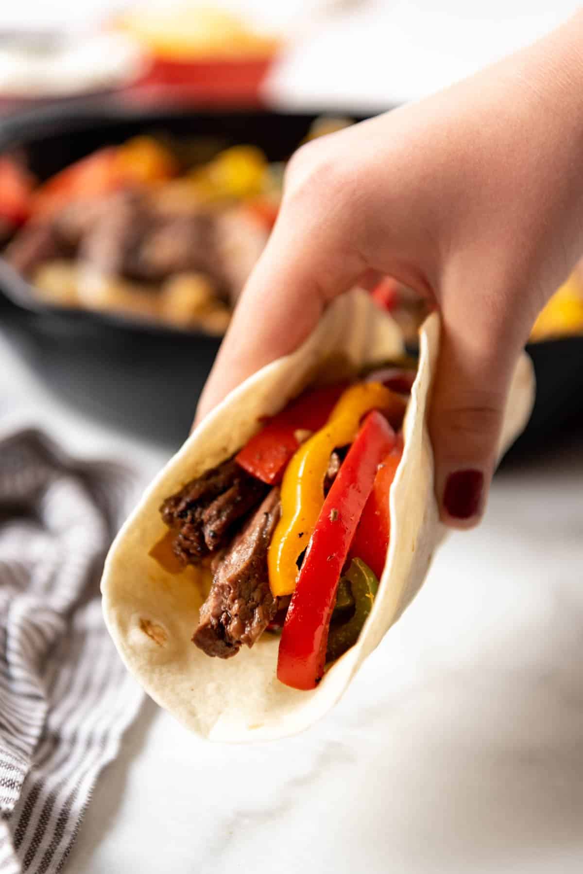 A hand lifting up a steak fajita with red and yellow peppers in a tortilla.