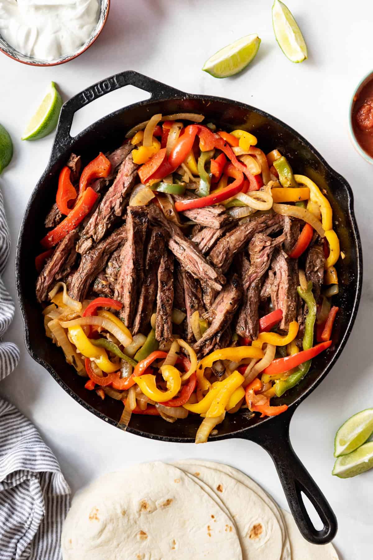 A cast iron skillet with steak fajita filling surrounded by tortillas, sour cream, and limes.