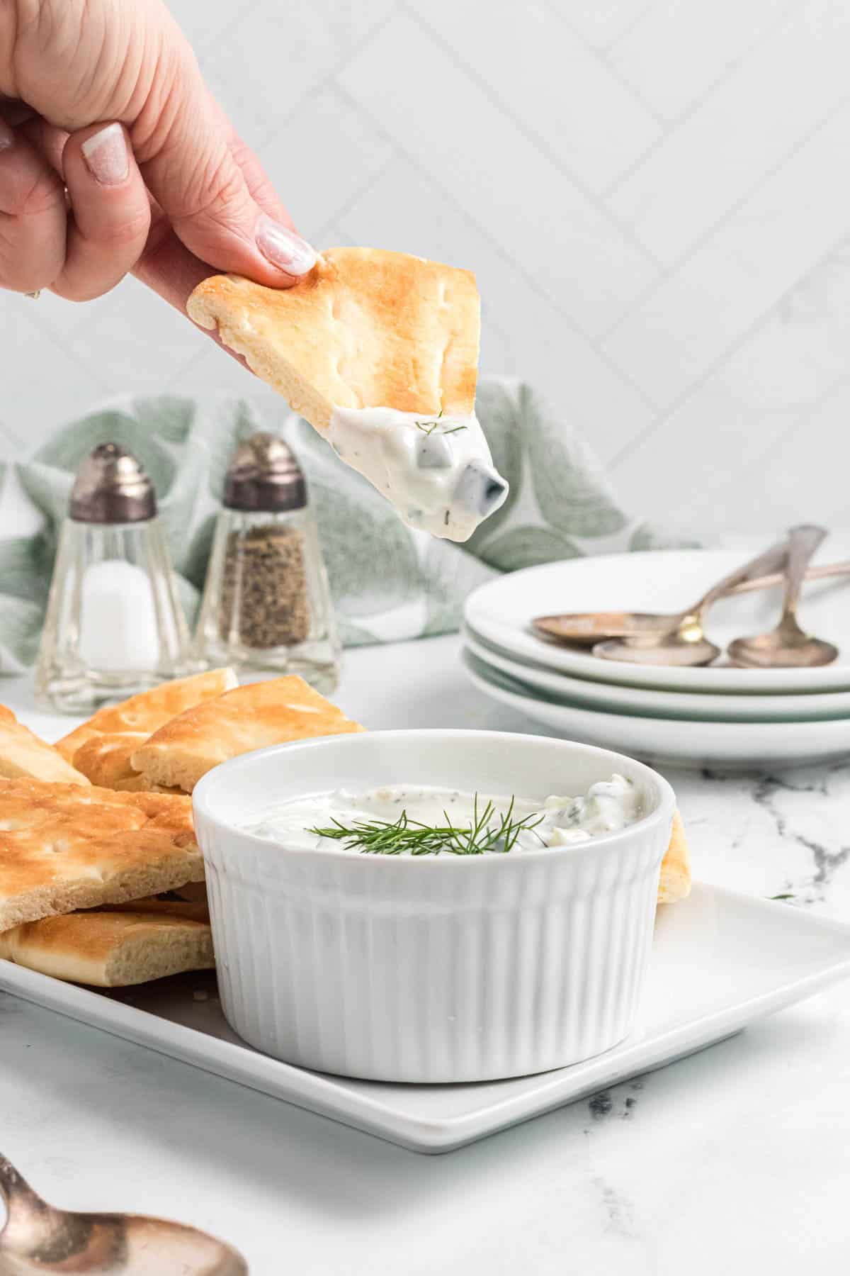 A hand holding a pita bread triangle to dip into tzatziki sauce.