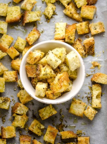 An overhead shot of a bunch of croutons spread across a sheet of parchment paper with a bowl full of croutons in the center