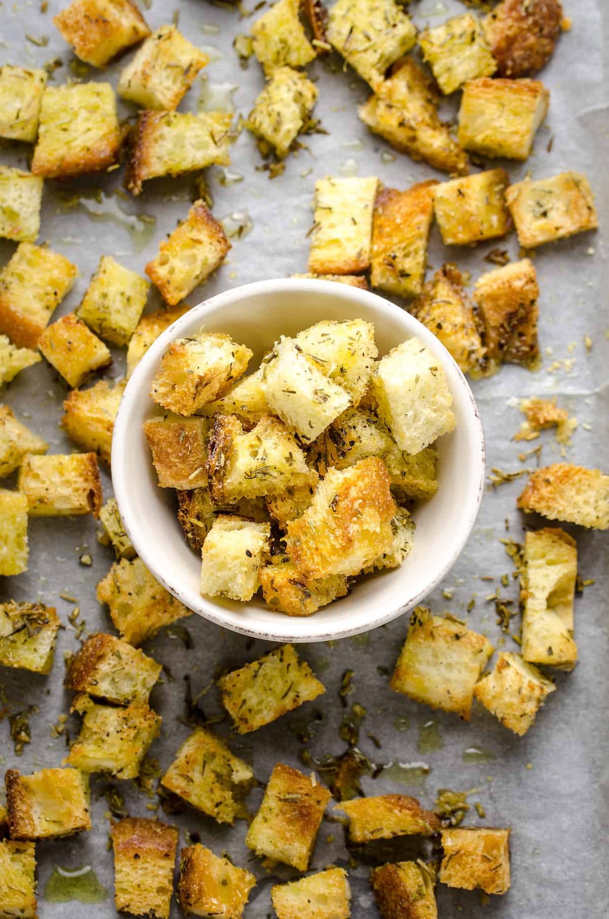 An overhead shot of a bunch of croutons spread across a sheet of parchment paper with a bowl full of croutons in the center