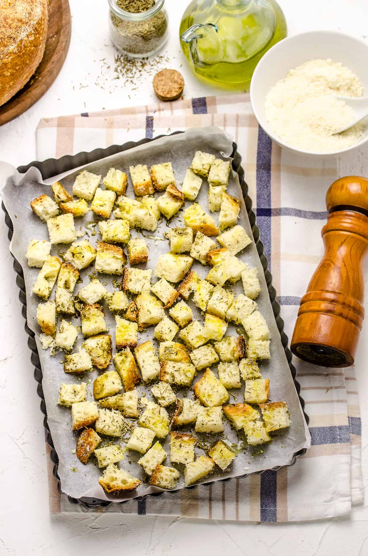 Seasoned cubes of bread on a lined baking sheet with a bowl of grated Parmesan beside them