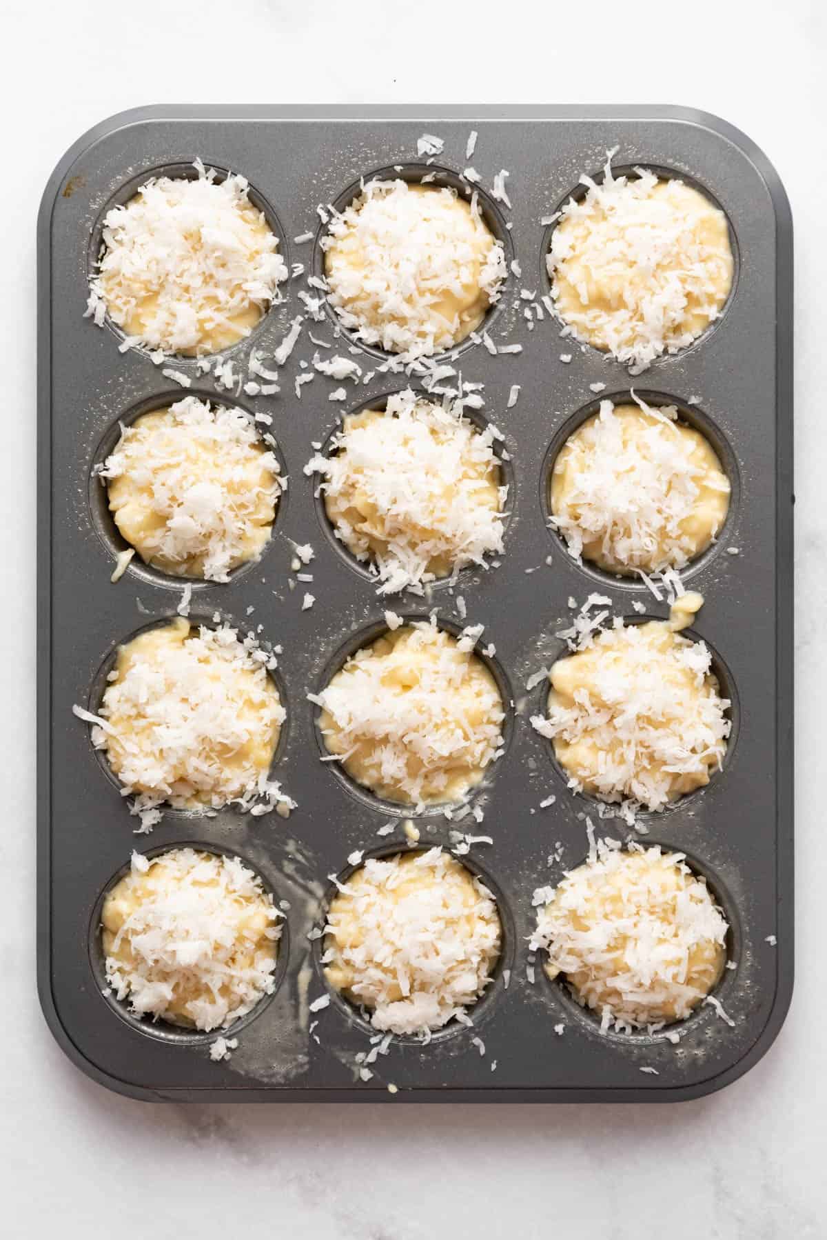 Unbaked pineapple muffin batter in a muffin pan with coconut sprinkled on top.
