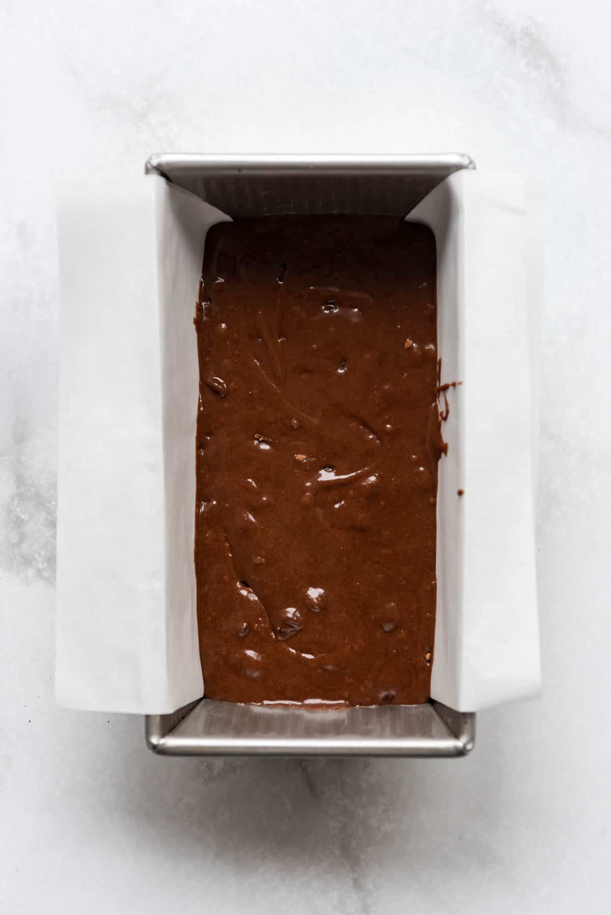 Brownie batter in a loaf pan with parchment paper sling.