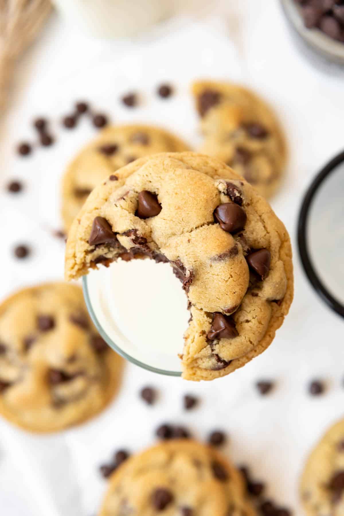 A chocolate chip cookie with a bite taken out of it balanced on the rim of a glass of milk.