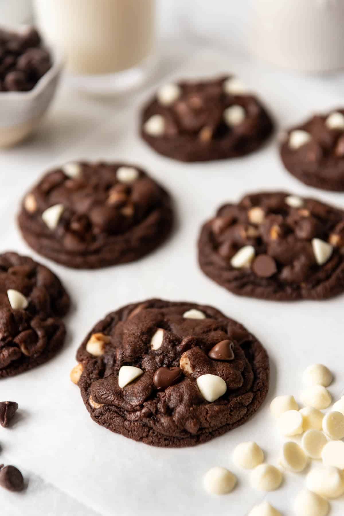 Six dark chocolate cookies on parchment paper next to white chocolate chips.