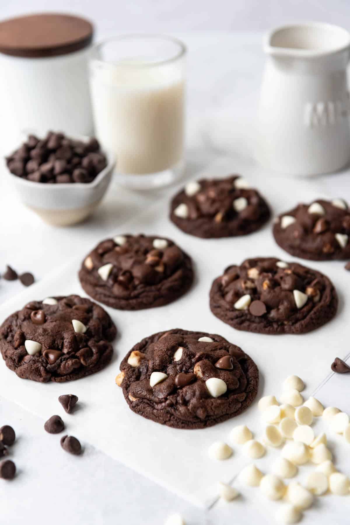 Six dark chocolate cookies with three types of chocolate chips in front of a glass of milk.