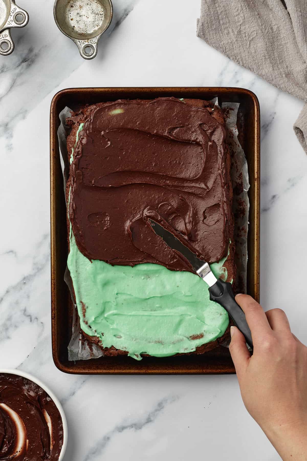 Chocolate frosting being spread onto a batch of brownies topped with mint frosting