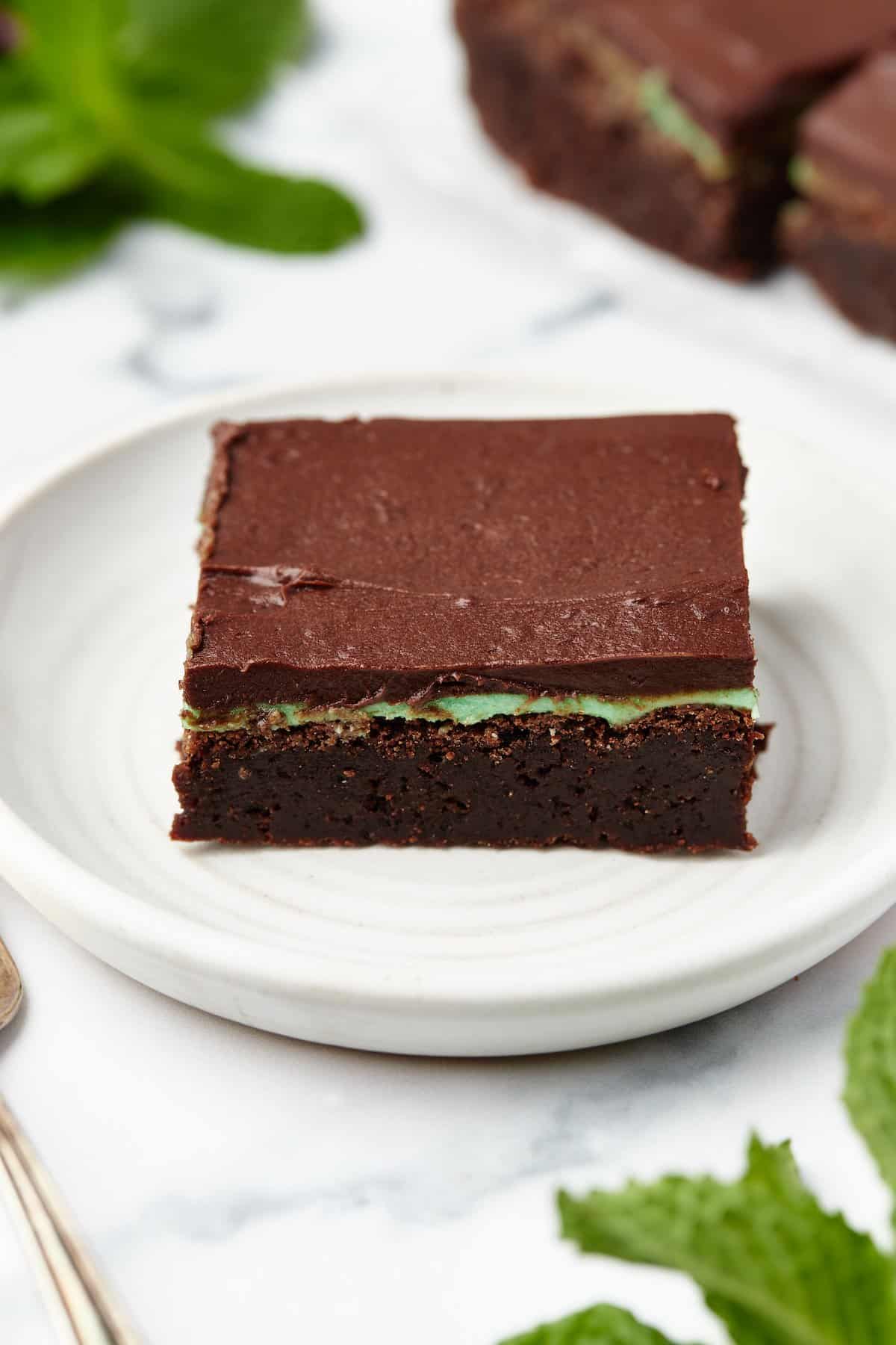 A peppermint brownie on a dessert plate with fresh mint leaves in front of it and behind it