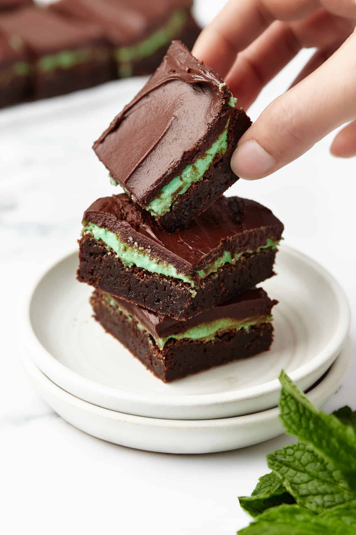 A stack of three peppermint brownies with a hand grabbing the one on top and a mint leaf in the foreground
