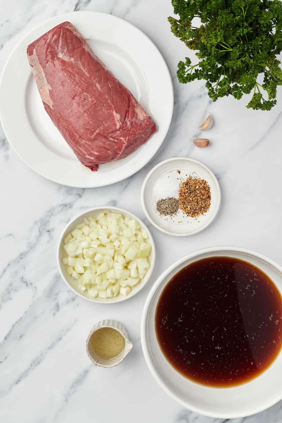 A raw chuck roast, steak seasoning, garlic cloves and the rest of the beef ingredients on a marble countertop