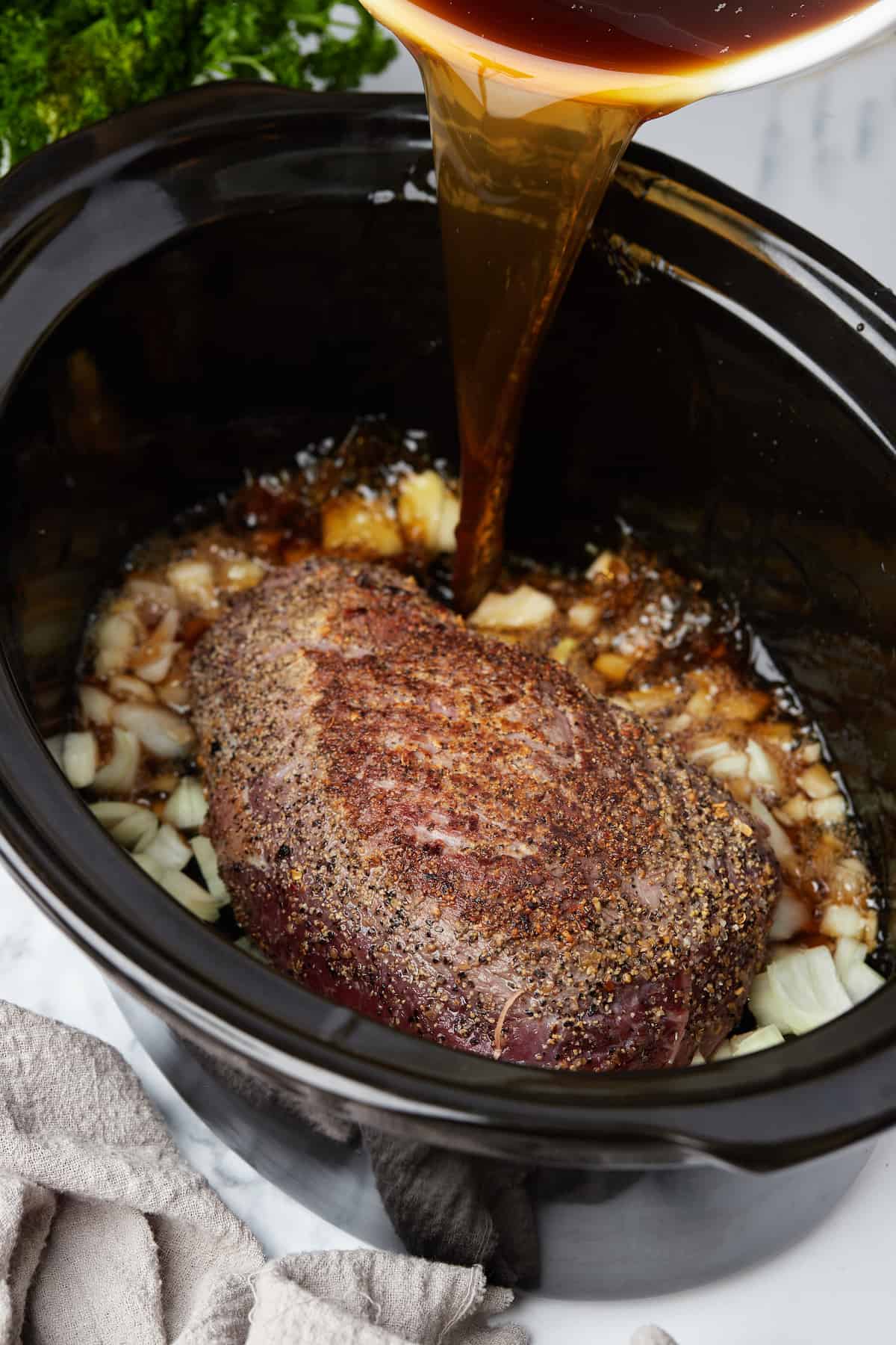 Beef broth being poured into a slow cooker filled with onions, garlic and beef