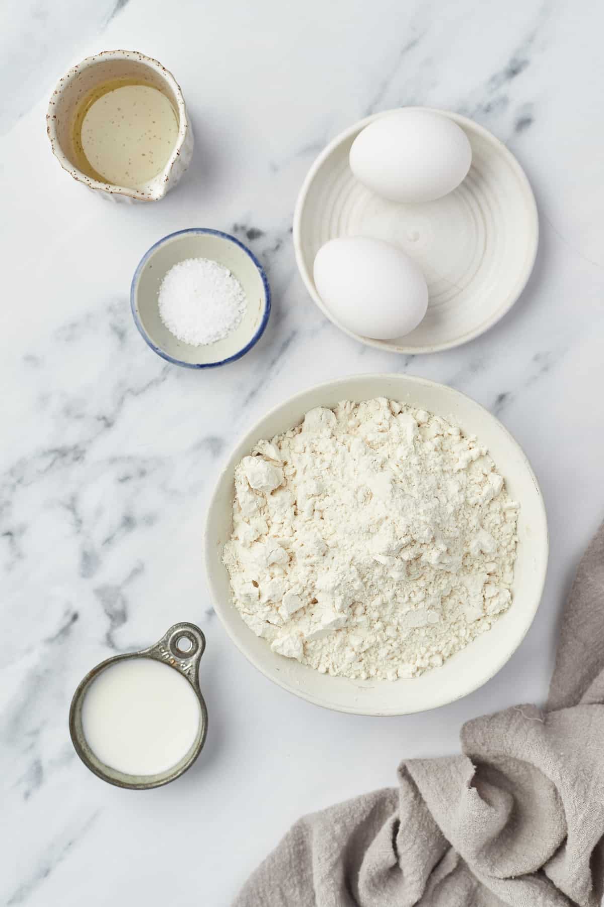 Two eggs, a bowl of flour and the rest of the egg noodle ingredients on a kitchen countertop