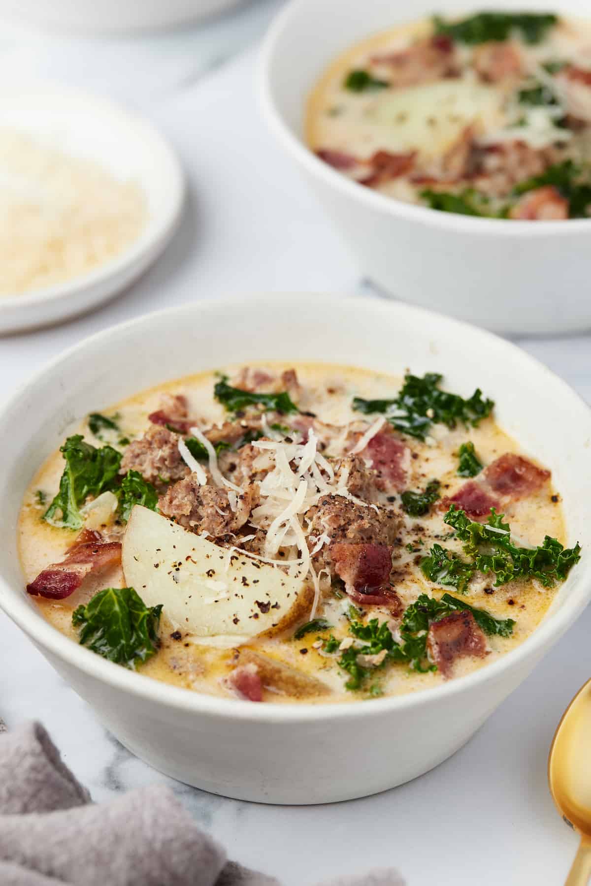 A bowl of Zuppa Toscana on a marble countertop with a plate of freshly grated cheese behind it