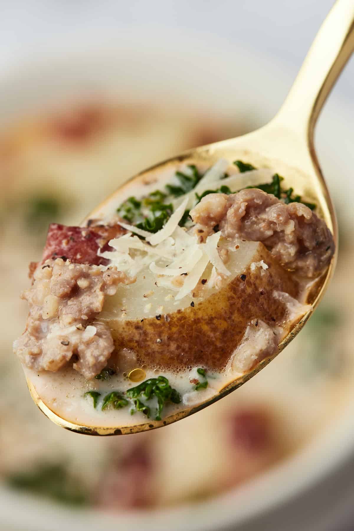 A mouthful of Zuppa Toscana soup on a golden spoon with a bowl full of soup behind it