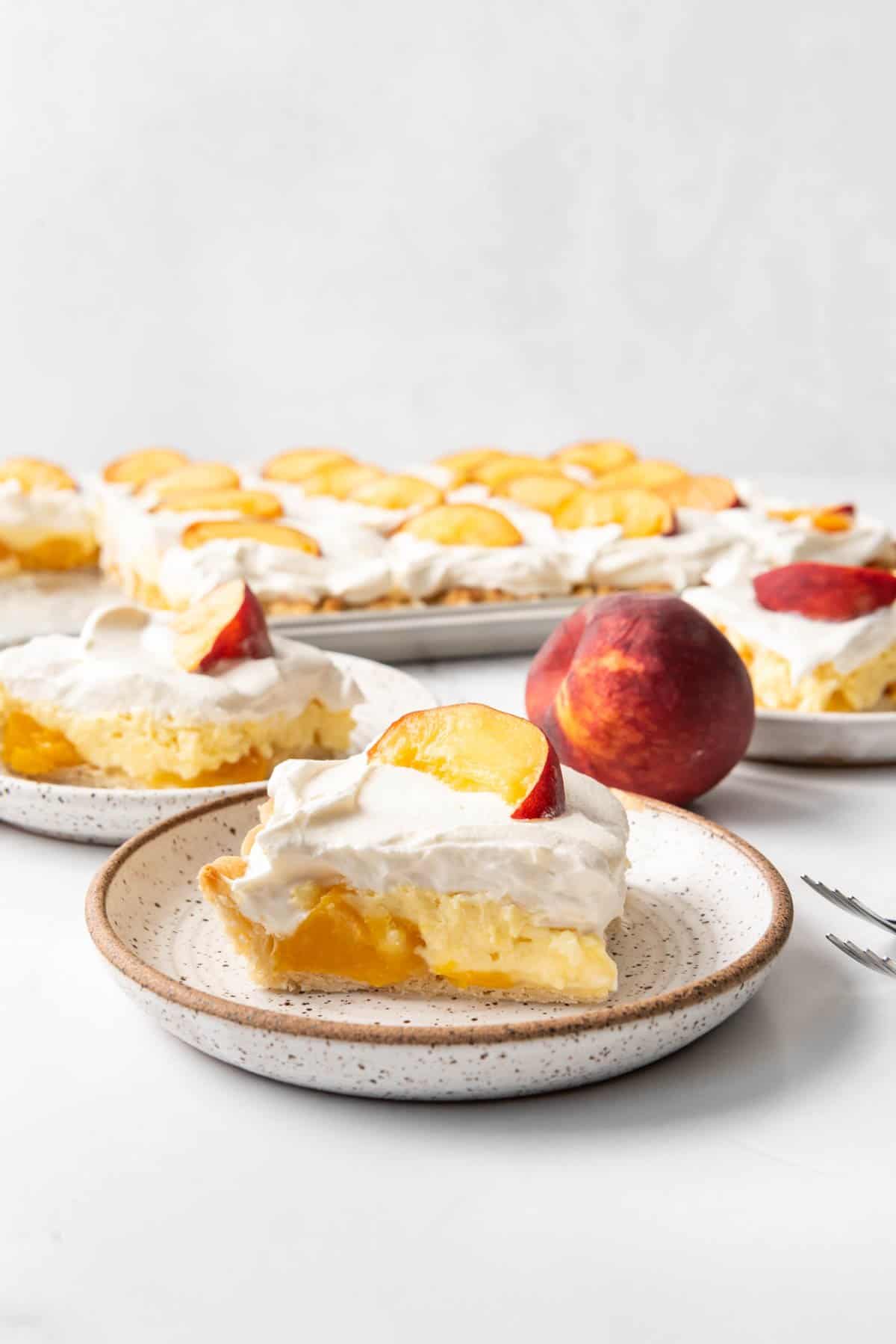 Slices of peaches and cream slab pie on plates next to a fresh peach.