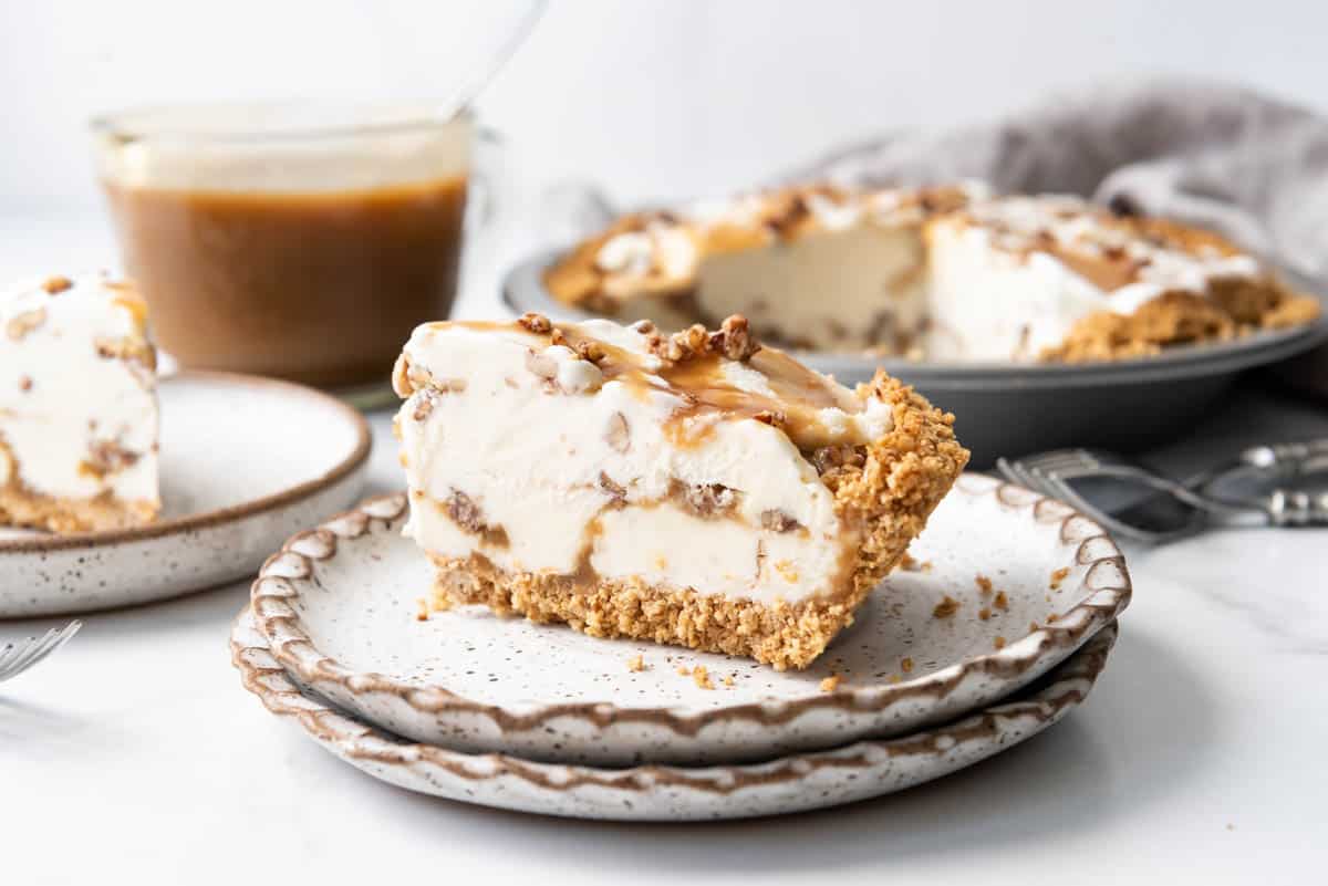 A slice of butter pecan ice cream pie on two stacked plates in front of a jar of butterscotch sauce.
