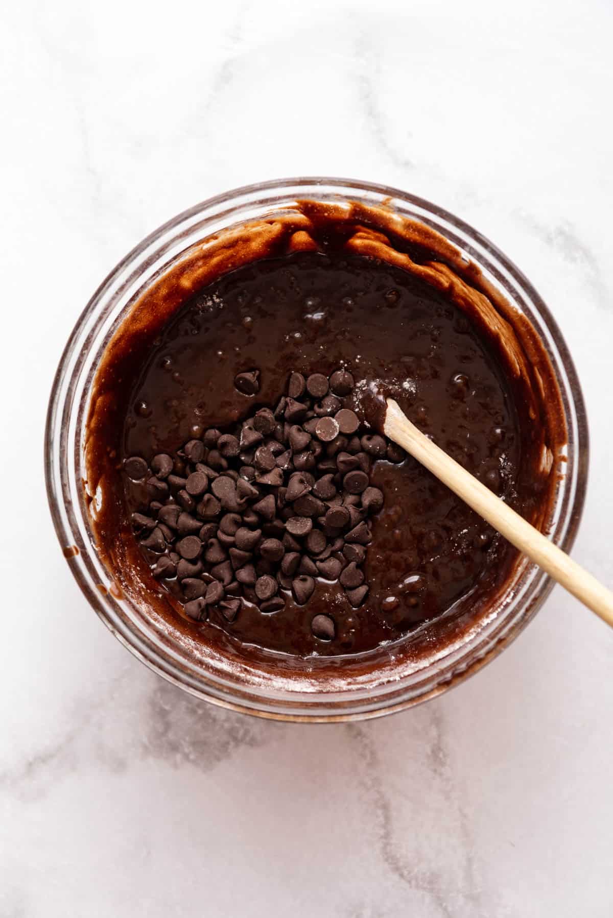 Adding chocolate chips to brownie batter in a glass bowl with a wooden spoon.