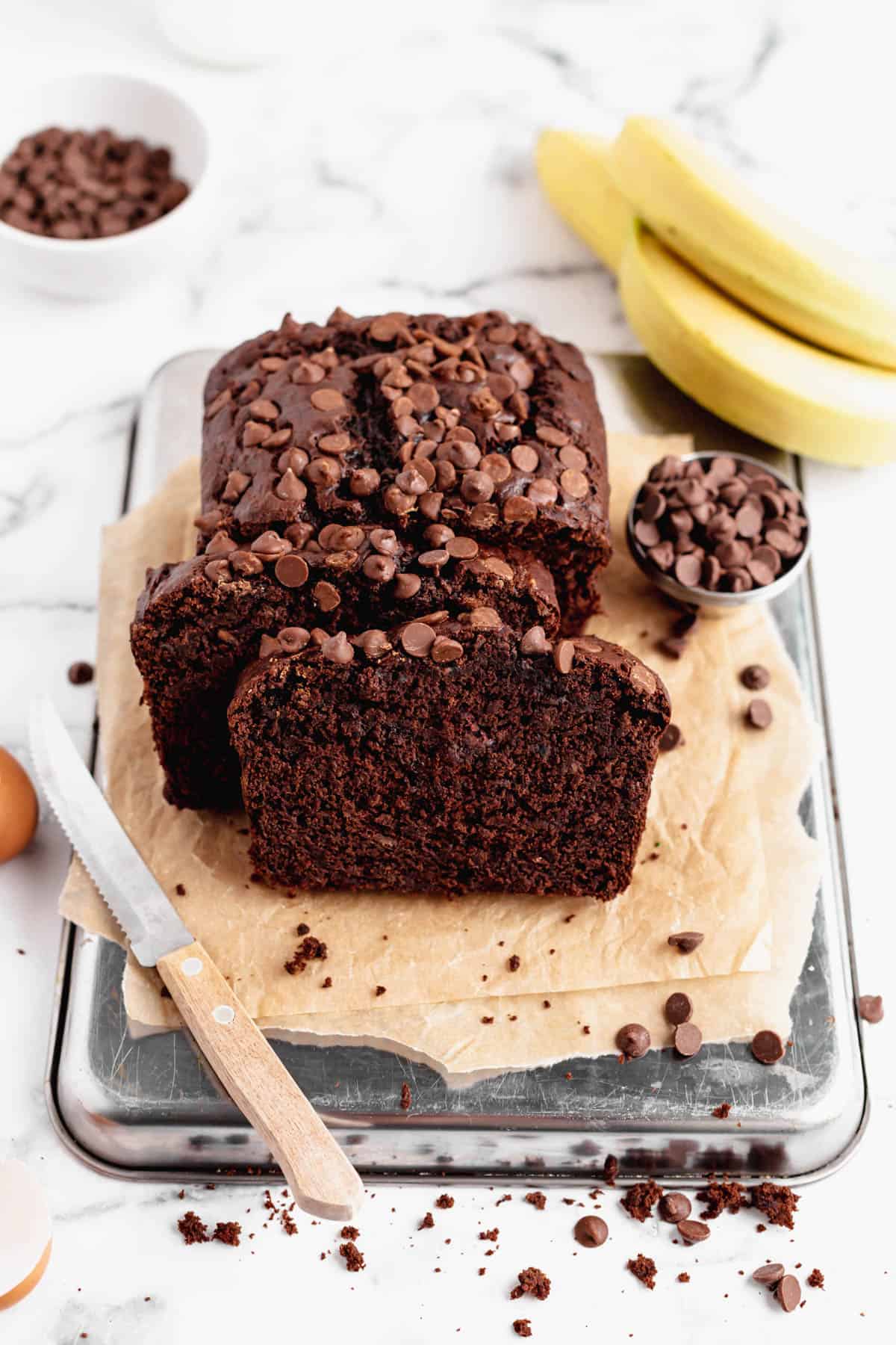 A chocolate banana bread loaf surrounded by chocolate chips, crumbs, a knife and bananas.
